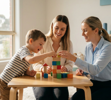 Parent and child smiling as they build a colorful block tower at a low table in a bright, sunlit playroom while a clinician provides gentle guidance nearby, creating a warm and hopeful therapy moment.