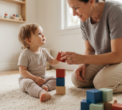 Warm candid close-up of a toddler and caregiver on a playroom floor making eye contact while stacking colorful blocks, with soft natural light and a blurred minimalist background.