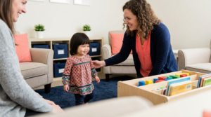 Sunlit therapy lobby where a therapist guides a toddler toward a child-sized play station, with organized bins and soft seating creating a warm, safe atmosphere.